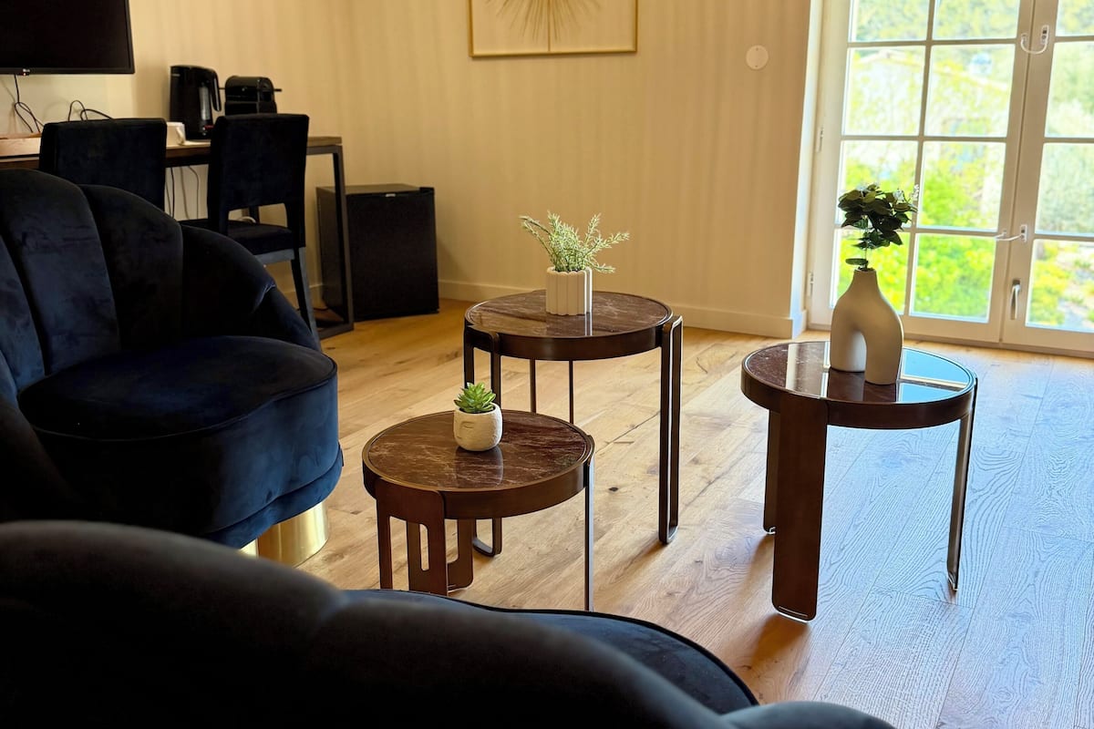 A living room with a dark blue sofa, a coffee table, a side table with a plant, and a large window with a view of greenery.