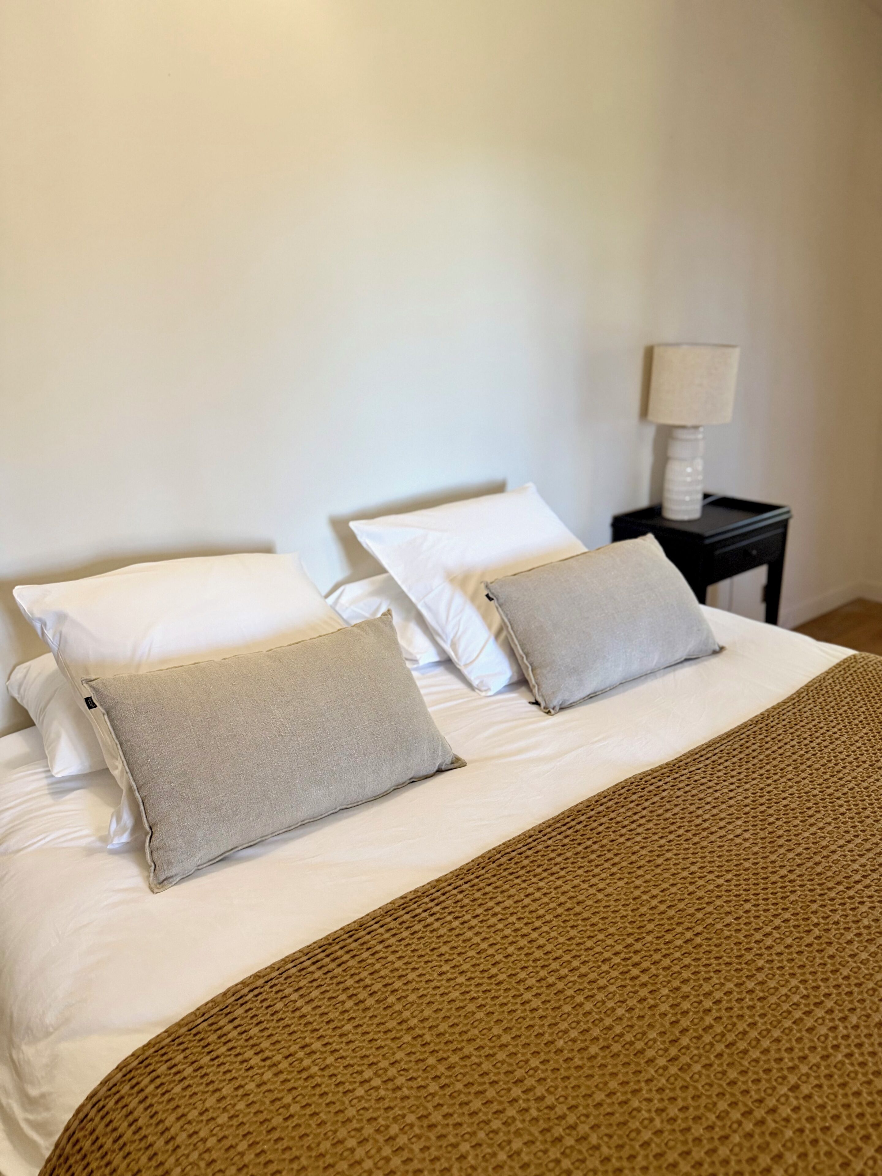 Ein Bett mit weißen und beigefarbenen Kissen, einer braunen, strukturierten Decke und einem kleinen Nachttisch mit Lampe.