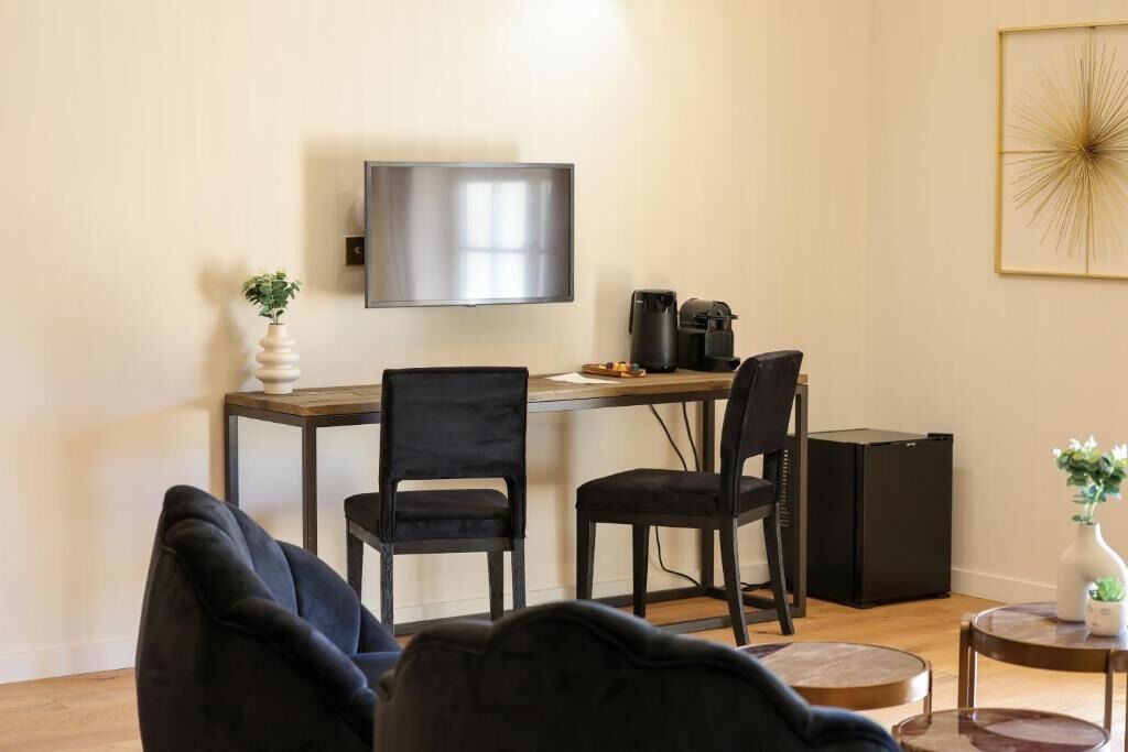 A modern living room with a flat-screen TV mounted on the wall, a wooden dining table with two chairs, a coffee machine, and a small side table with a plant.