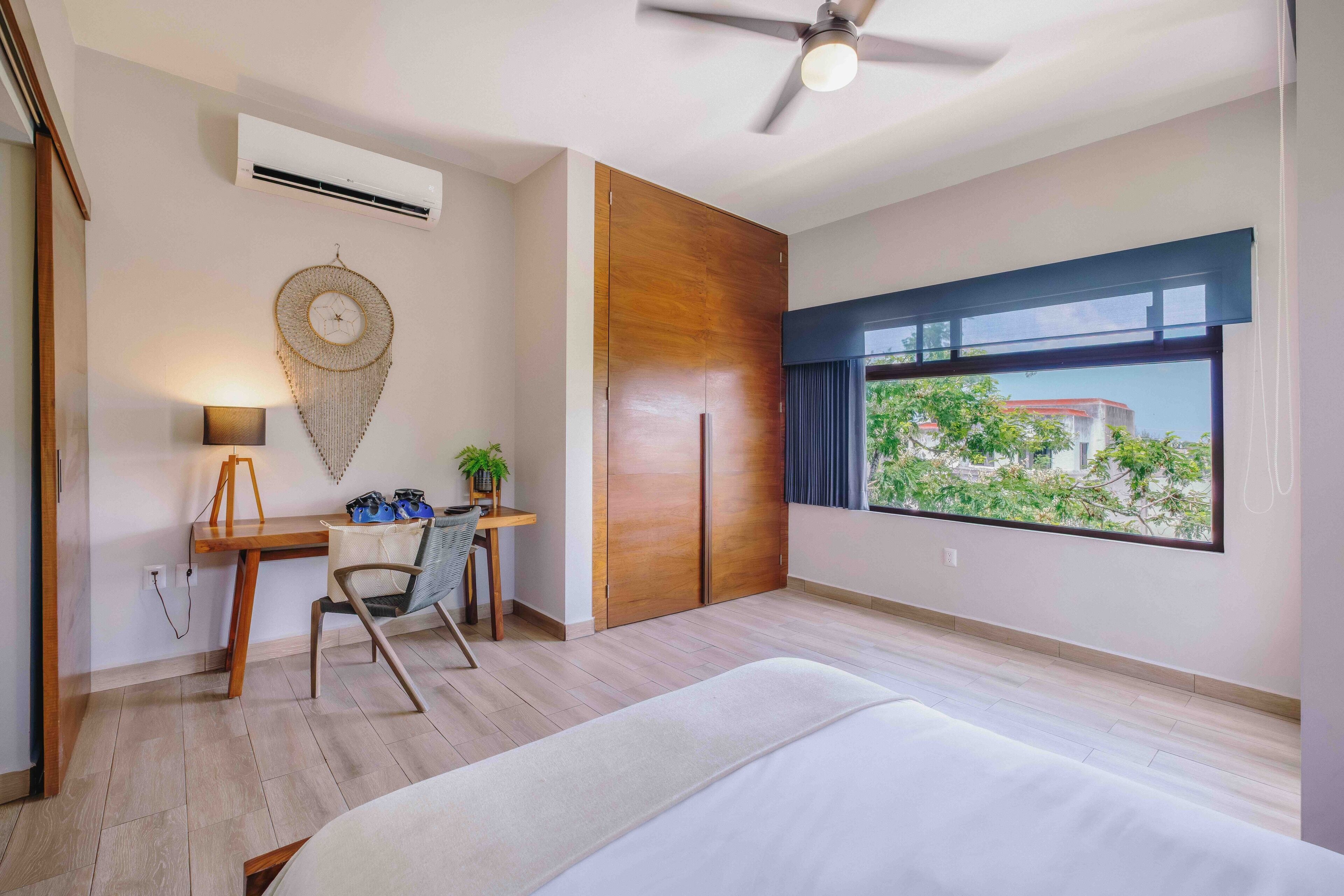 A bedroom with a bed, a desk with a chair, a ceiling fan, an air conditioner, and a window with a view of greenery.