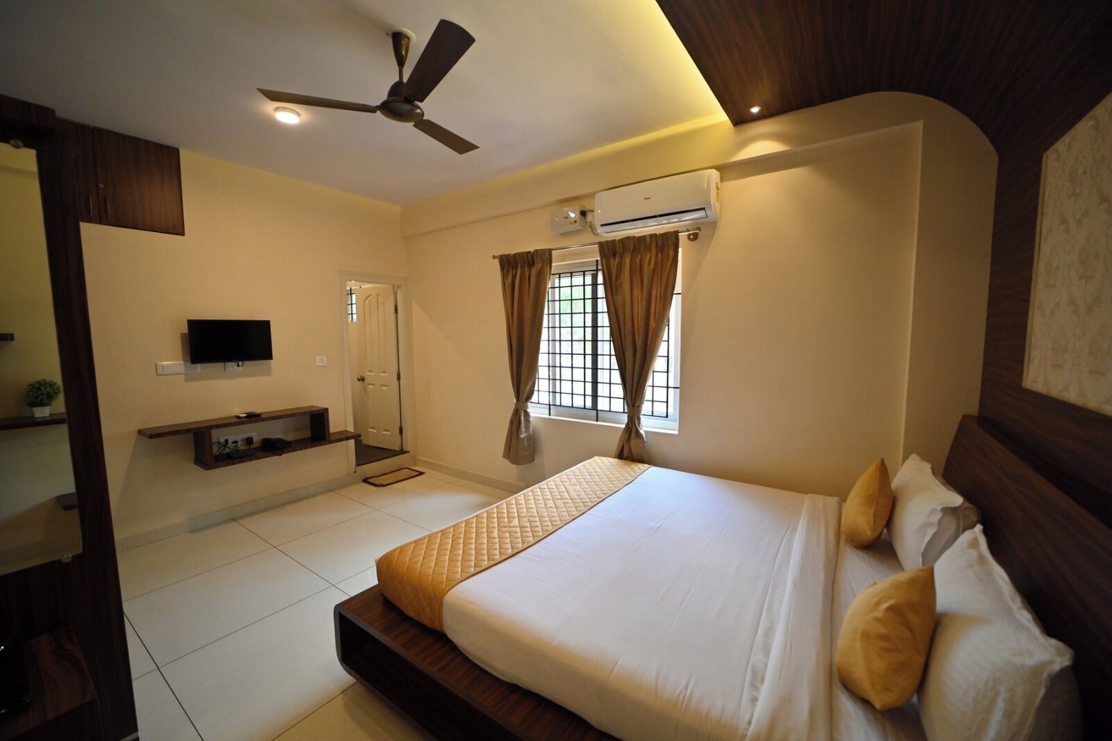 A bedroom with a bed, a ceiling fan, an air conditioning unit, a TV, and a window with curtains.