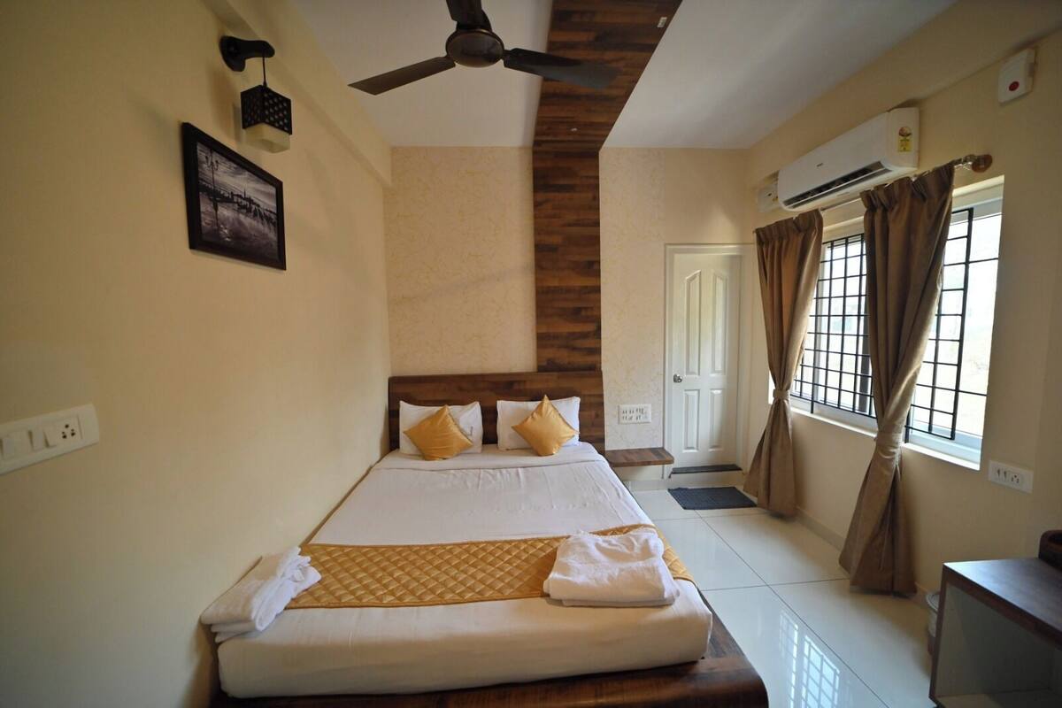 A hotel room with a bed, ceiling fan, air conditioning unit, and a window with curtains.