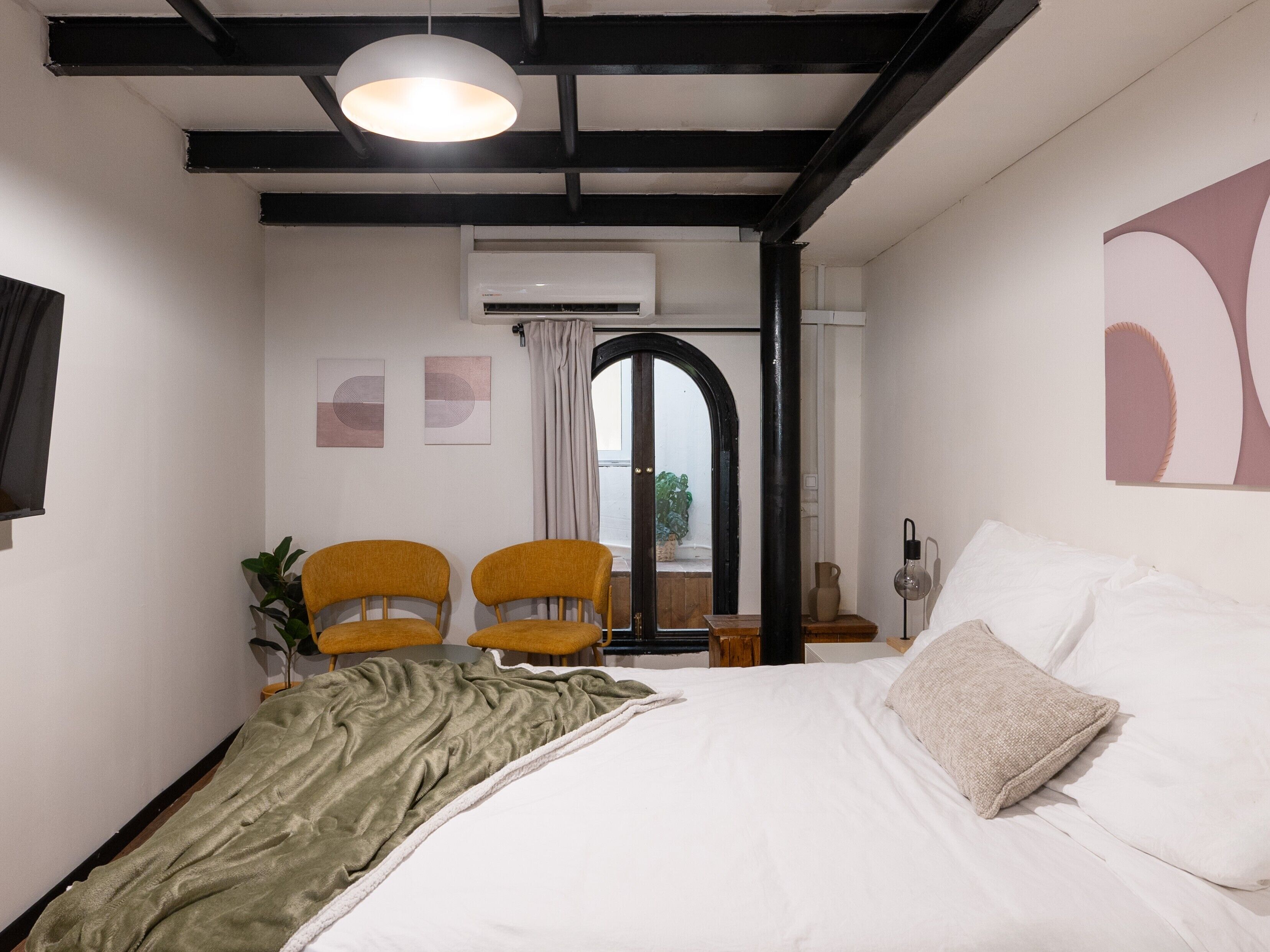 A bedroom with a bed, two chairs, a window with curtains, an air conditioning unit, and a ceiling light.