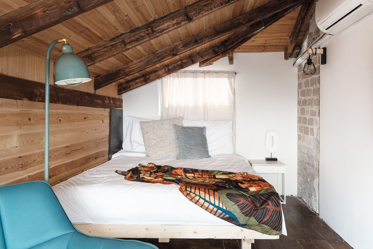 Un dormitorio con techo de madera, una cama con ropa blanca, una silla azul y una mesita de noche con lámpara.