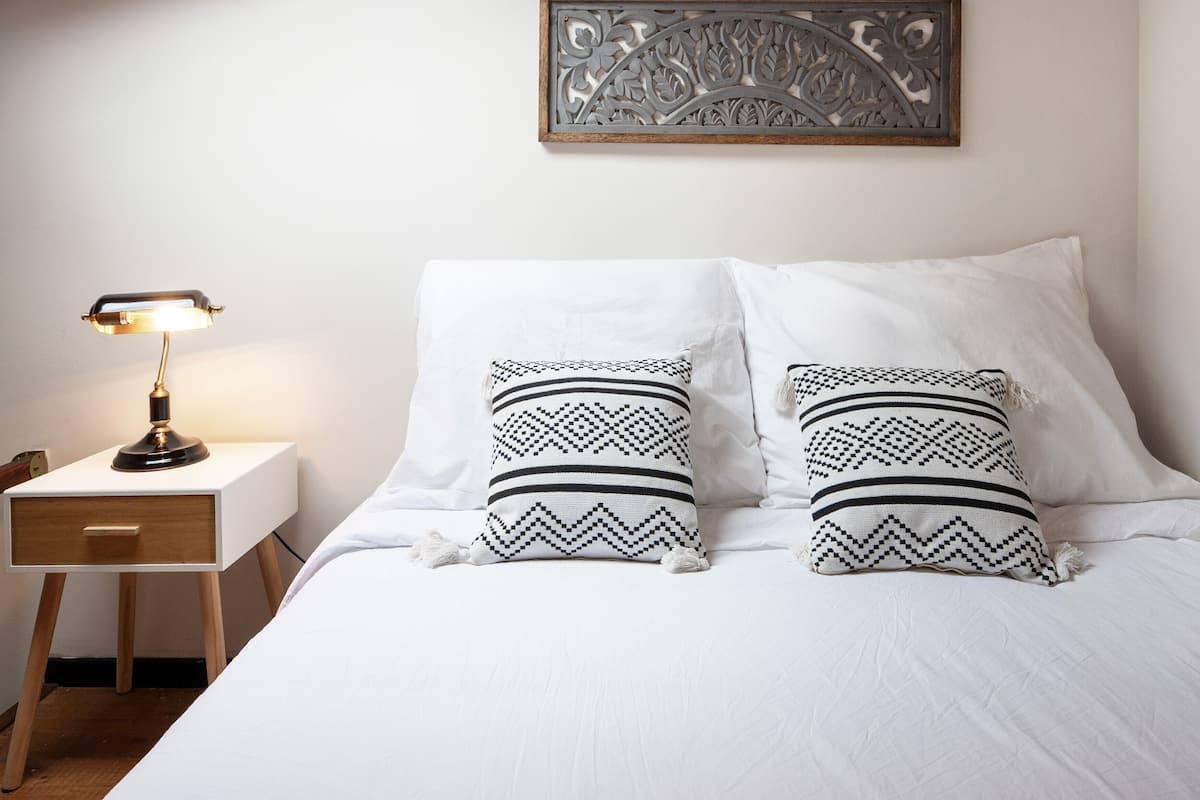 Una cama bien hecha con dos almohadas estampadas, una lámpara de noche y un adorno en la pared.