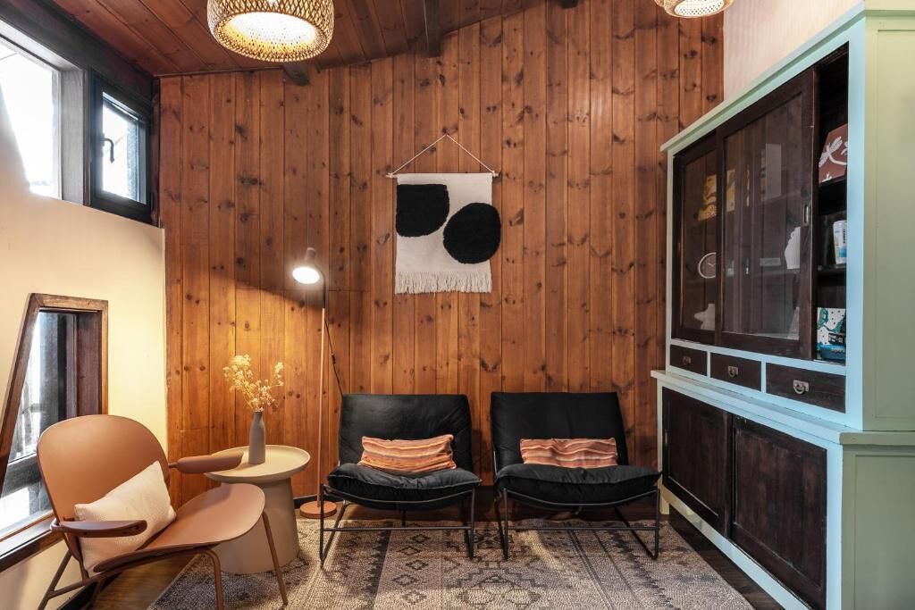A room with wooden panel walls, two black chairs with cushions, a small round table with a vase, and a cabinet with glass doors.