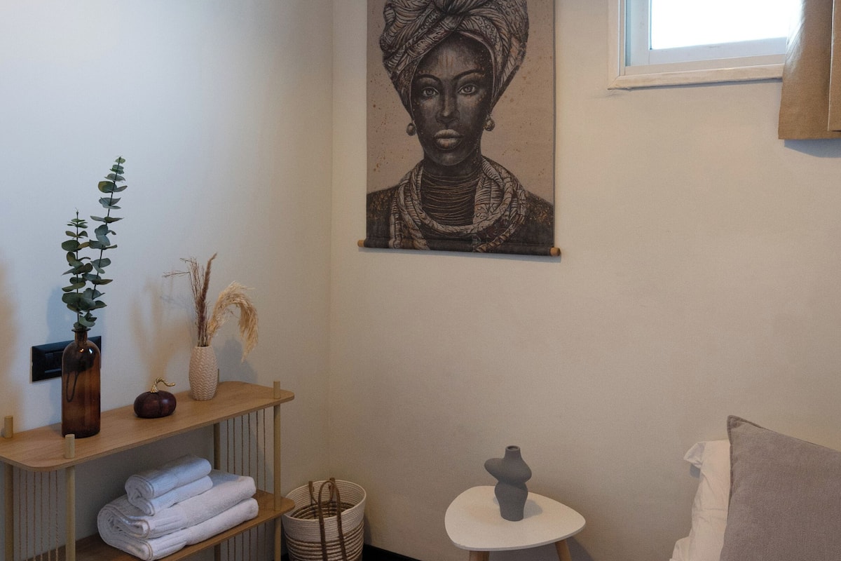 Un dormitorio con piso de madera, una cama con ropa blanca, una mesita de noche con lámpara, un estante con toallas dobladas y una obra de arte en la pared.