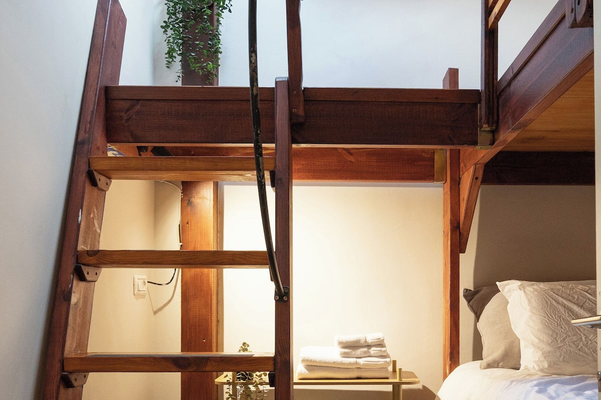 Una habitación pequeña con una cama nido, una escalera de madera, un estante con toallas y un reloj pequeño.