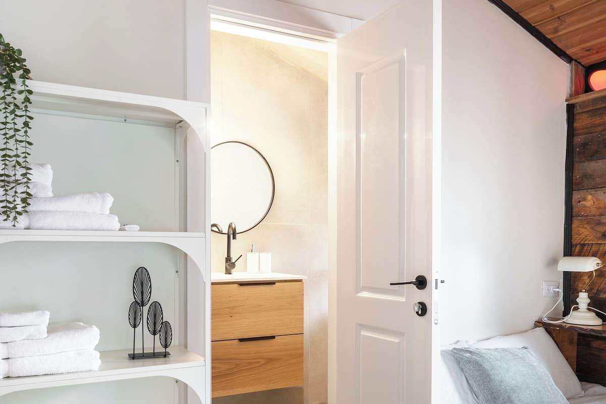 Un dormitorio con techo de madera, una puerta blanca, una cama con ropa de cama blanca, una mesita de noche con lámpara y un estante blanco con toallas y un objeto decorativo.