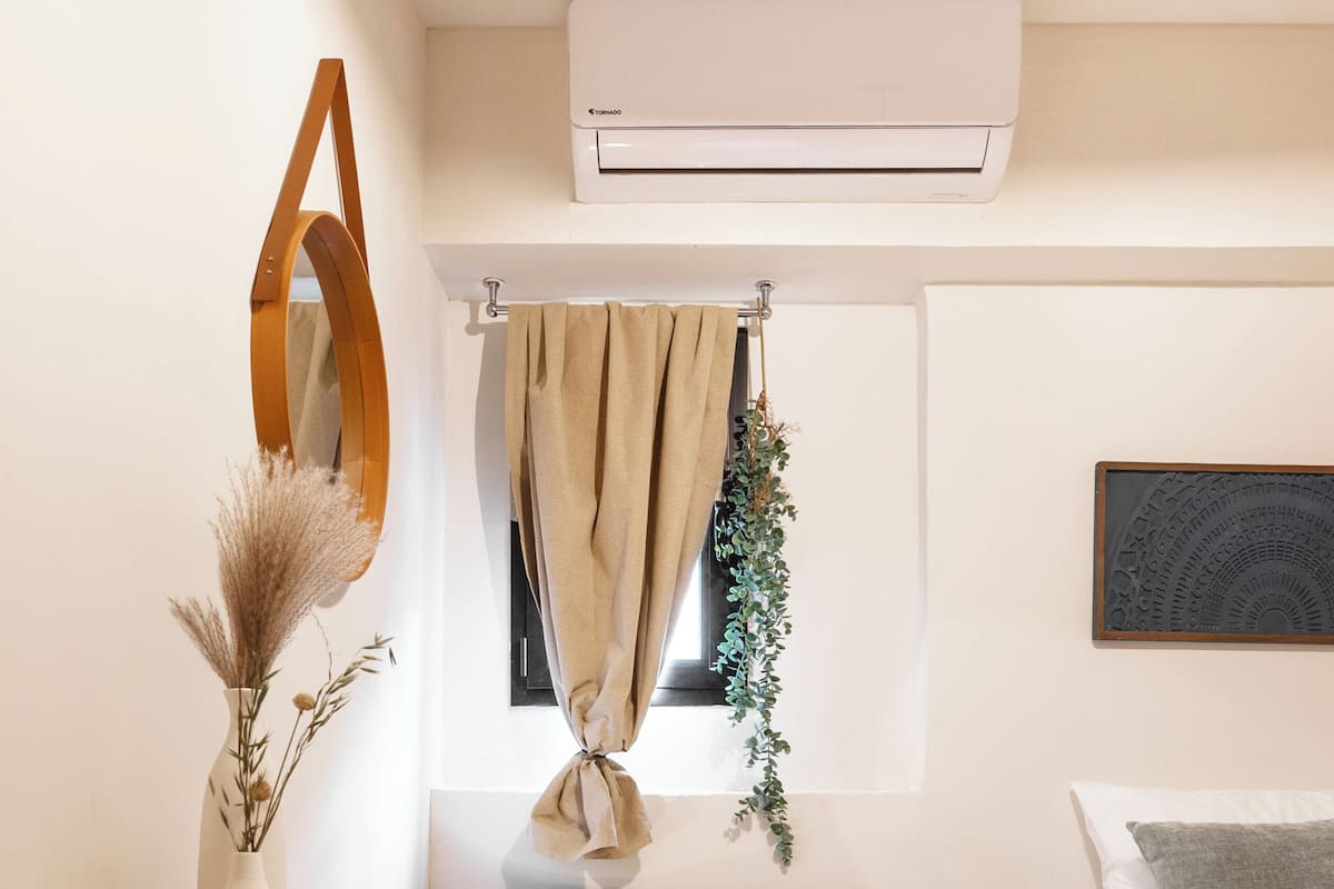 Un dormitorio moderno con cama, una silla, una mesita, un estante con toallas, una ventana con cortinas y un equipo de aire acondicionado.