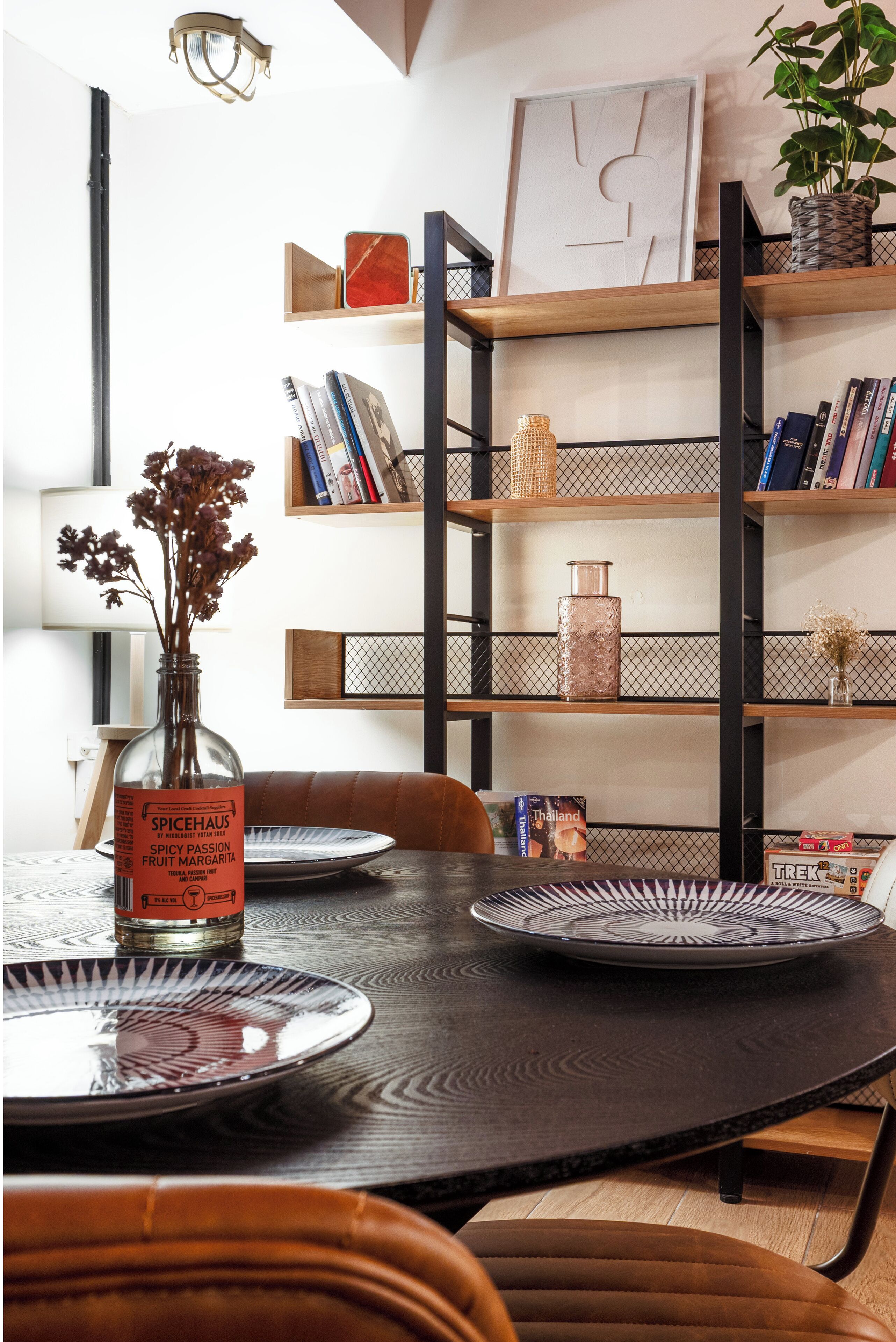 A dining area with a round table, chairs, a vase with dried flowers, a bookshelf, and a lamp.