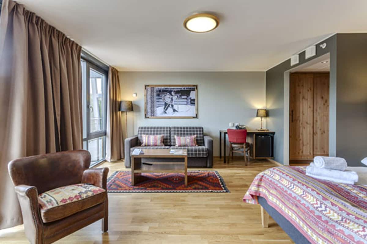 Una habitación de hotel con cama, sofá, silla, mesita y ventana con cortinas.