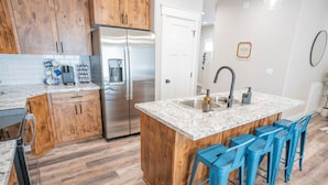 A kitchen with wooden cabinets, a stainless steel refrigerator, a marble countertop, and blue bar stools. - Ashton Escape - Newly built townhome near Yellowstone/Grand Tetons/Island Park (Ashton)