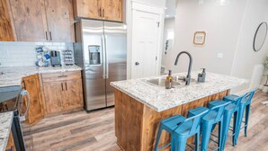 A kitchen with wooden cabinets, a stainless steel refrigerator, a marble countertop, and blue bar stools. - Ashton Escape - Newly built townhome near Yellowstone/Grand Tetons/Island Park (Ashton)