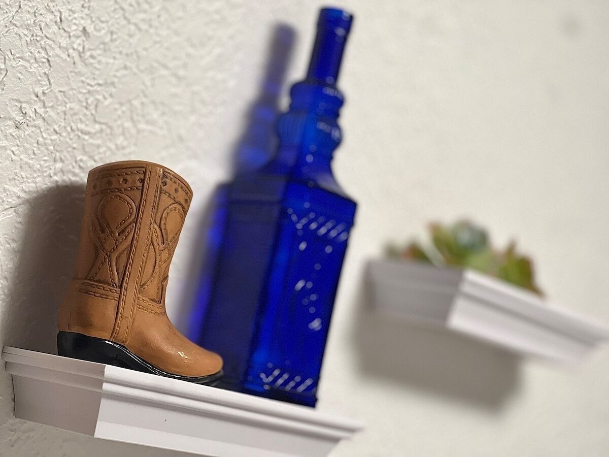 Una bota vaquera de madera y una botella azul sobre estantes blancos frente a una pared con textura.