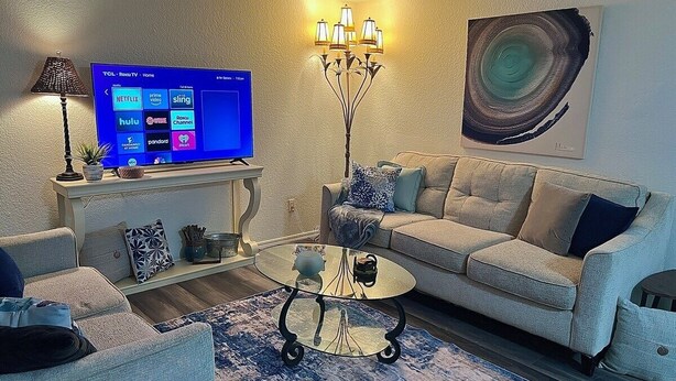 Una sala de estar con sofá, sillones, una mesa de centro, televisor de pantalla plana, lámpara y un cuadro en la pared.
