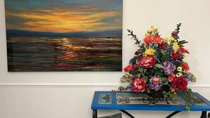 Un cuadro de un atardecer sobre el agua en una pared, una mesa con un florero y un mantel azul.