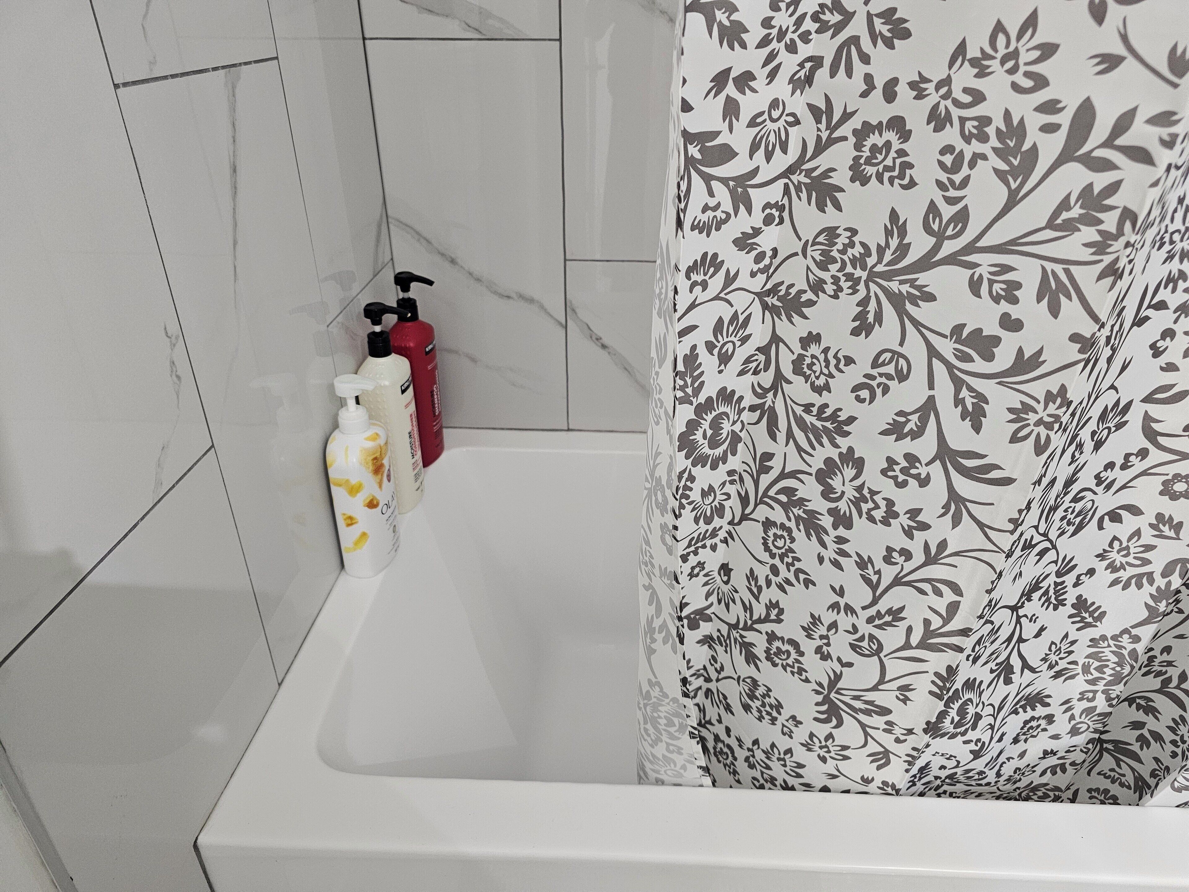 Et badeværelse med et hvidt badekar, en blomstret bruseforhæng og to flasker shampoo og balsam.