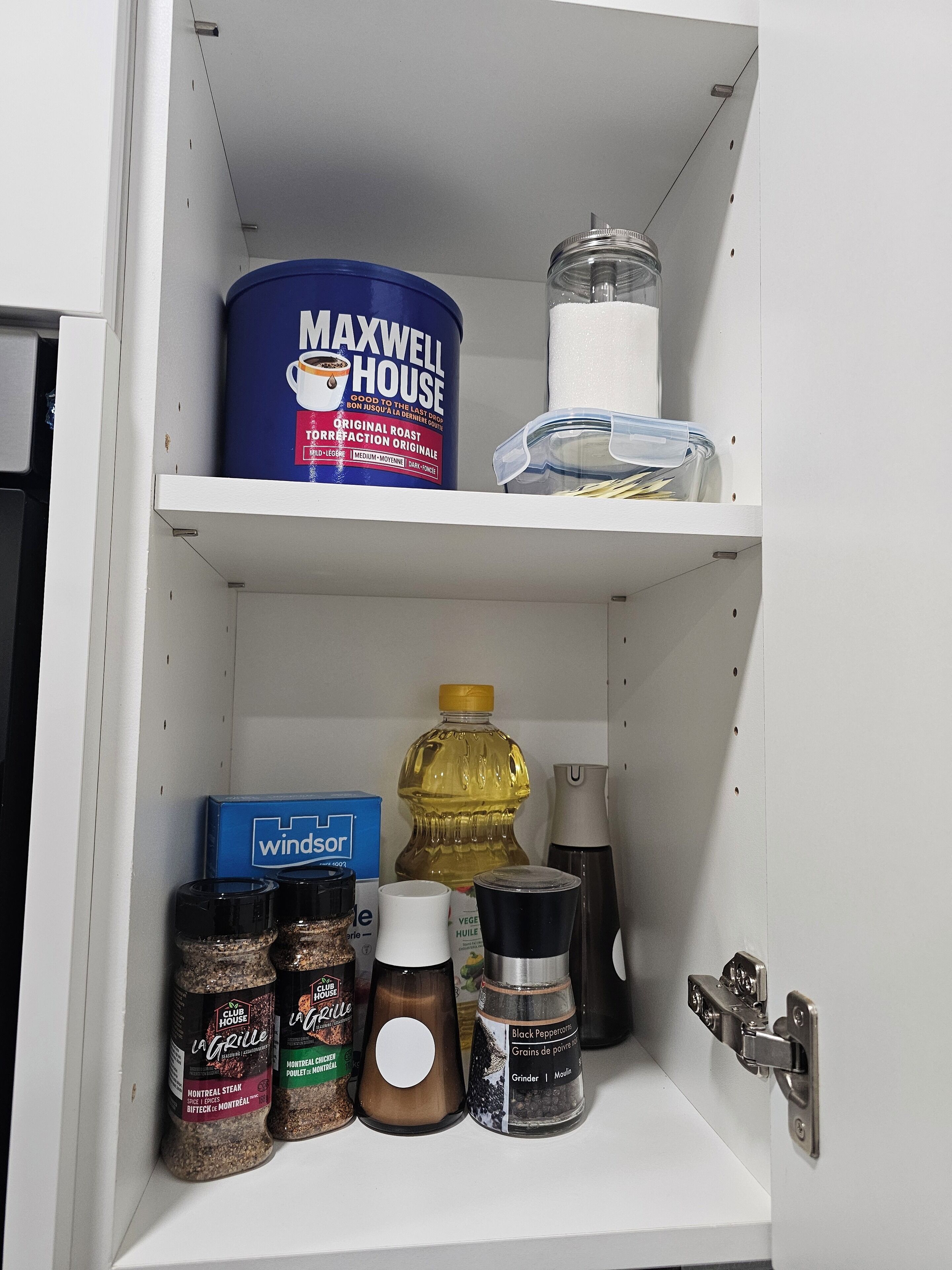 A kitchen cabinet with a blue canister labeled 'MAXWELL HOUSE', a clear jar, a bottle of oil, and various spice containers.