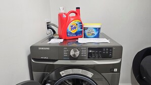 Un lave-linge Samsung à chargement frontal, une bouteille de lessive rouge et une boîte de lessive bleue posées sur le dessus.