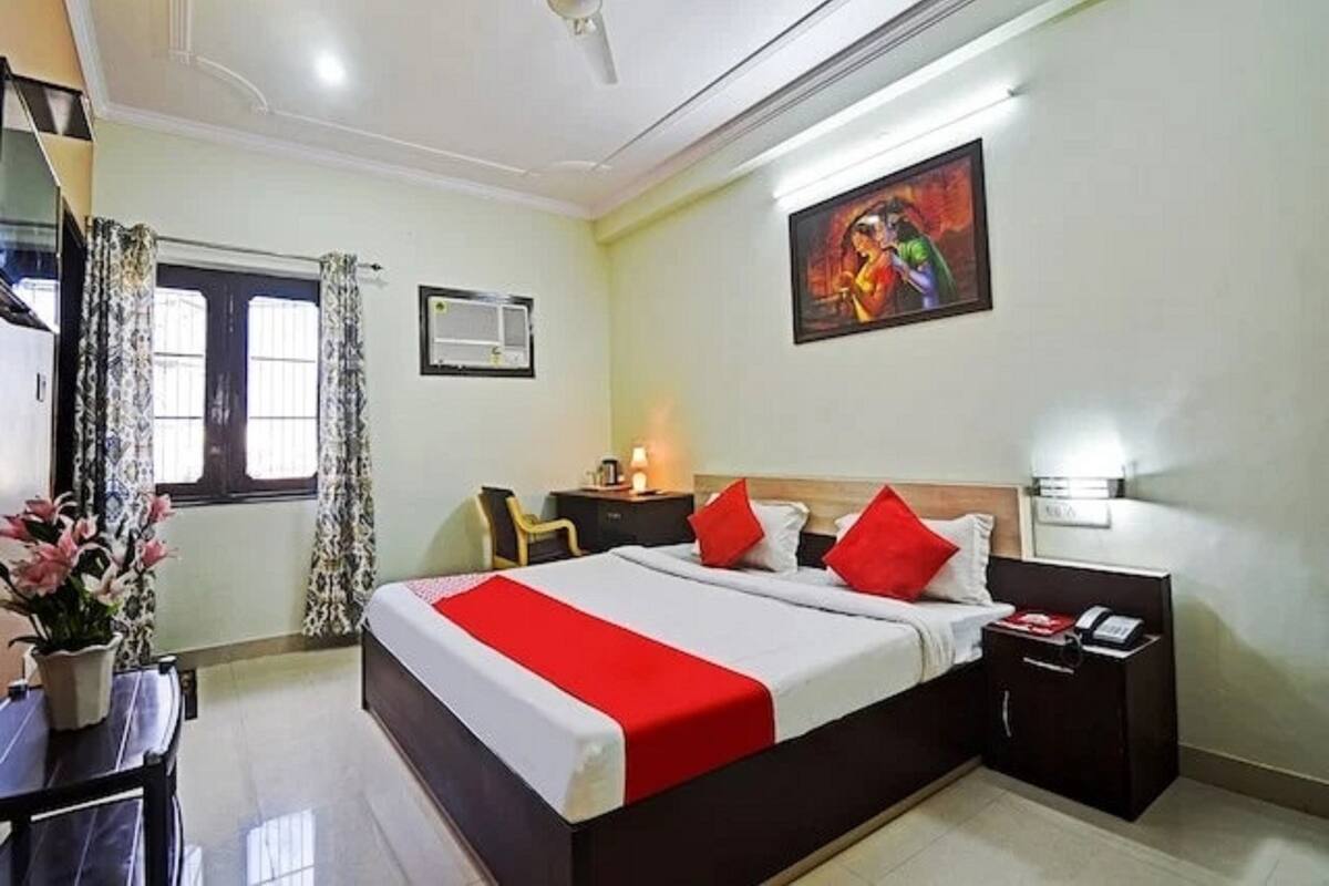 A hotel room with a bed, bedside tables, a desk, a chair, a window with curtains, an air conditioning unit, and a painting on the wall.