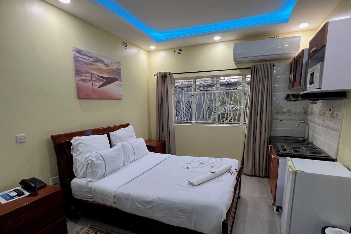 A hotel room with a bed, a small kitchenette, a window with curtains, and a patterned rug.