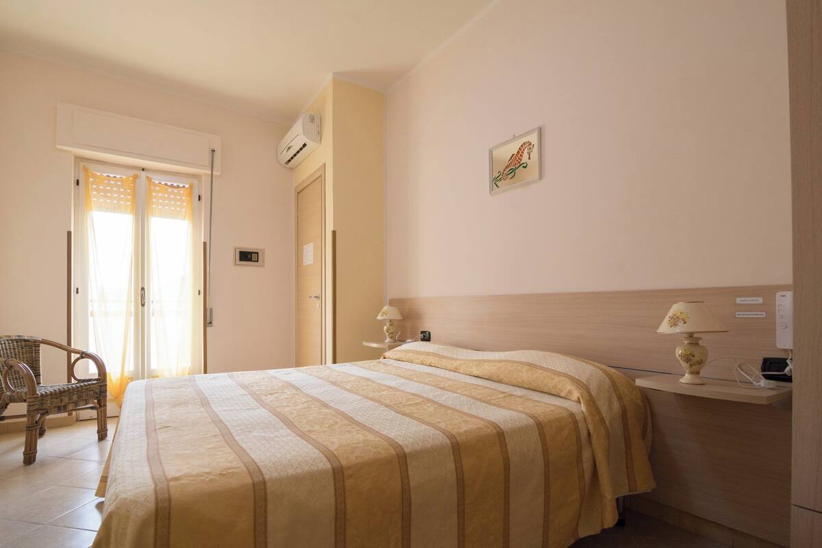 A hotel room with a bed, a chair, a nightstand, and a wall-mounted air conditioner.