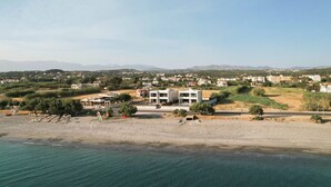 A coastal property with a modern two-story building, a sandy beach, and a view of a town in the distance. - Gemini Villas | Villa Siesta (Μάλεμε)