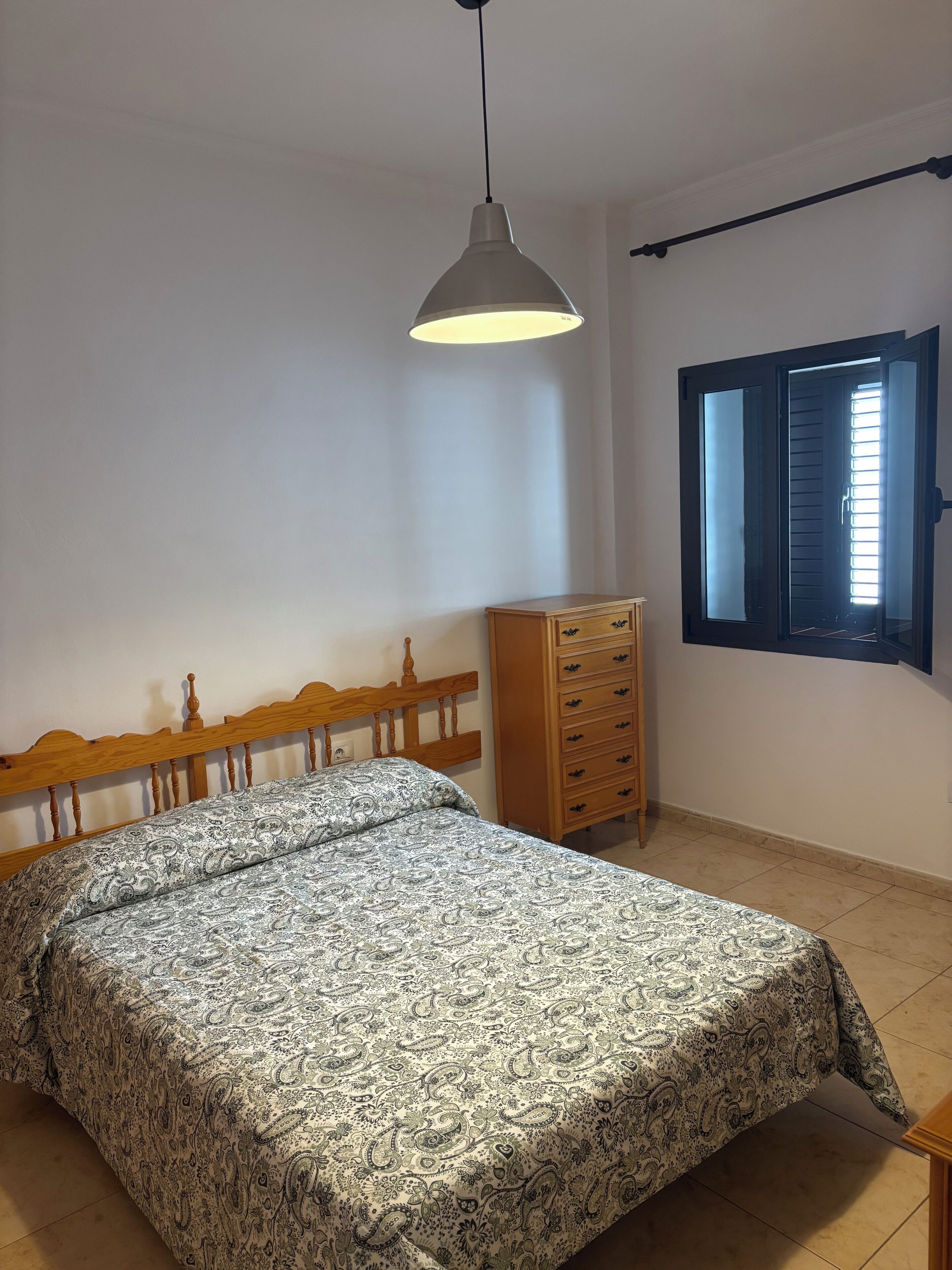 Une chambre à coucher comprenant un lit, une commode, une fenêtre avec des stores et un luminaire suspendu.