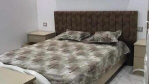 A bedroom with a bed, a nightstand, a dresser, and a wall-mounted light fixture. - Appartement Kelibia (Kerkouane)