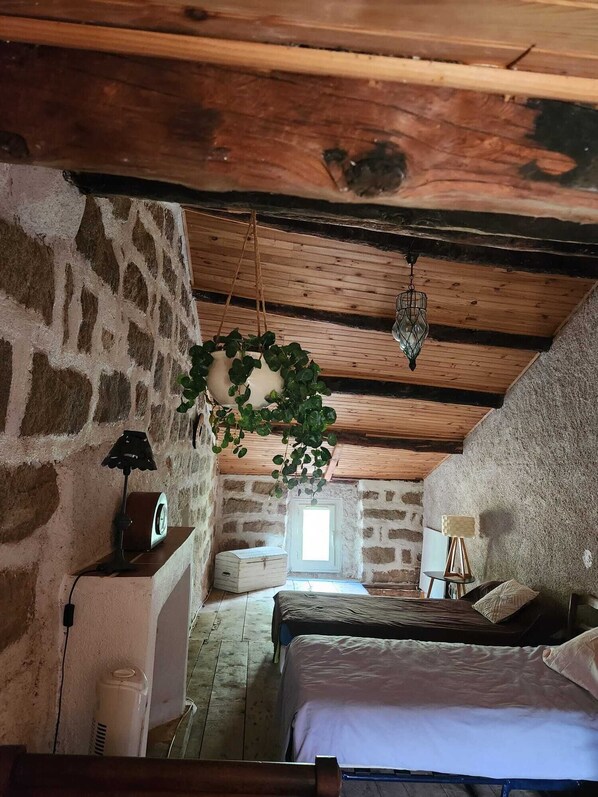 A rustic bedroom with exposed wooden beams, a stone wall, a hanging plant, a bed, a desk, and a window.