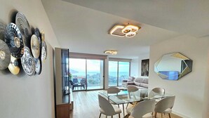 A spacious living area with a glass dining table, modern chairs, and a large window offering a city view.
