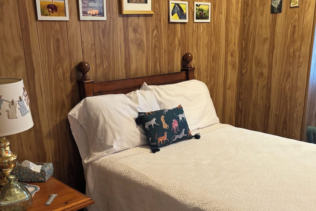 Une chambre à coucher avec une tête de lit en bois, un lit recouvert de draps blancs, un coussin décoratif, une table de chevet avec une lampe et des tableaux encadrés accrochés au mur.