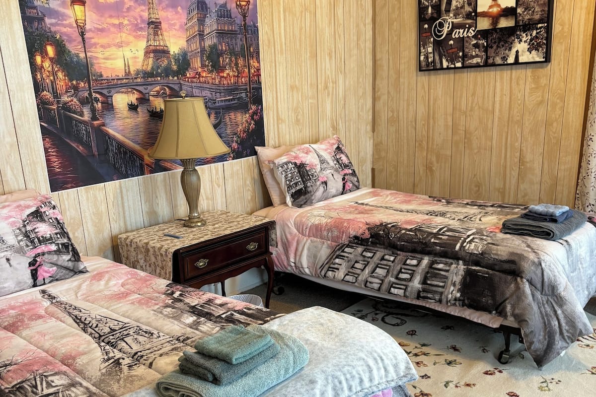 Une chambre avec deux lits, une table de chevet, une lampe et un grand tableau sur le thème de Paris accroché au mur.
