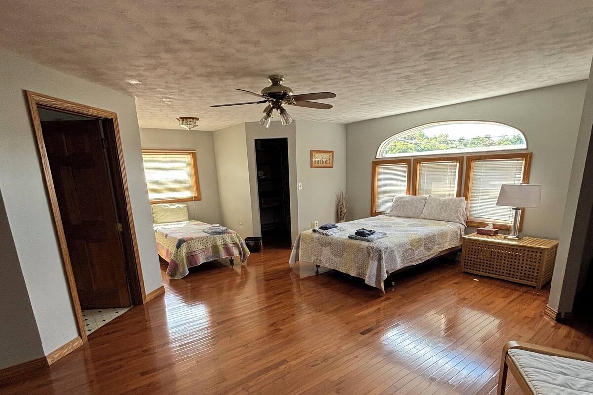 Une pièce spacieuse dotée d’un plancher en bois, de deux lits avec des têtes de lit, d’un ventilateur de plafond et d’une grande fenêtre avec des stores.