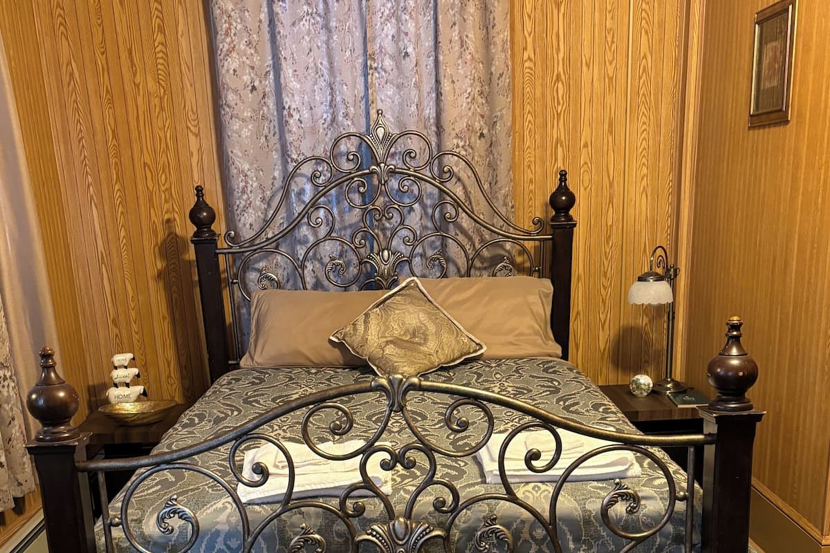 Un lit avec un cadre en métal orné, une couvre-lit à motifs et un coussin décoratif. Il y a une table de chevet avec une lampe et un petit objet décoratif. La pièce est dotée de lambris en bois et d’un rideau.