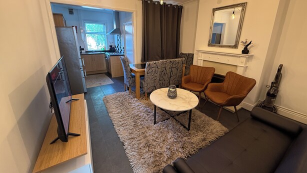 Una sala de estar moderna con un sofá, sillones, una mesa de centro y un televisor. Se observa una cocina al fondo con una ventana y un comedor.