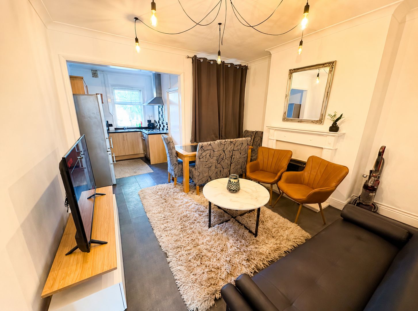 A compact living space with a sofa, chairs, a small table, a TV, and a kitchen in the background.