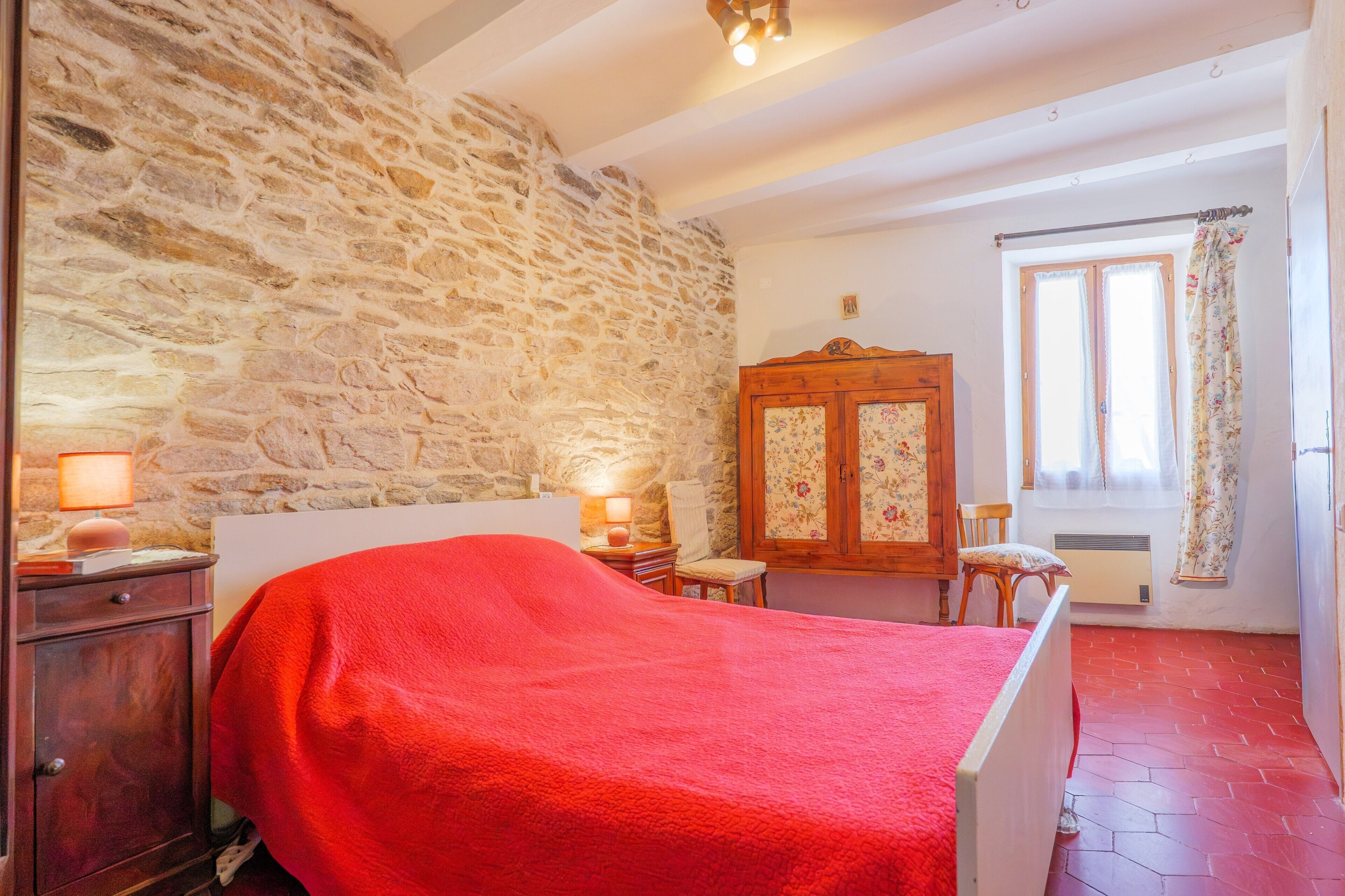 A bedroom with a stone wall, a bed with a red cover, a wooden wardrobe, a chair, and a window with curtains.