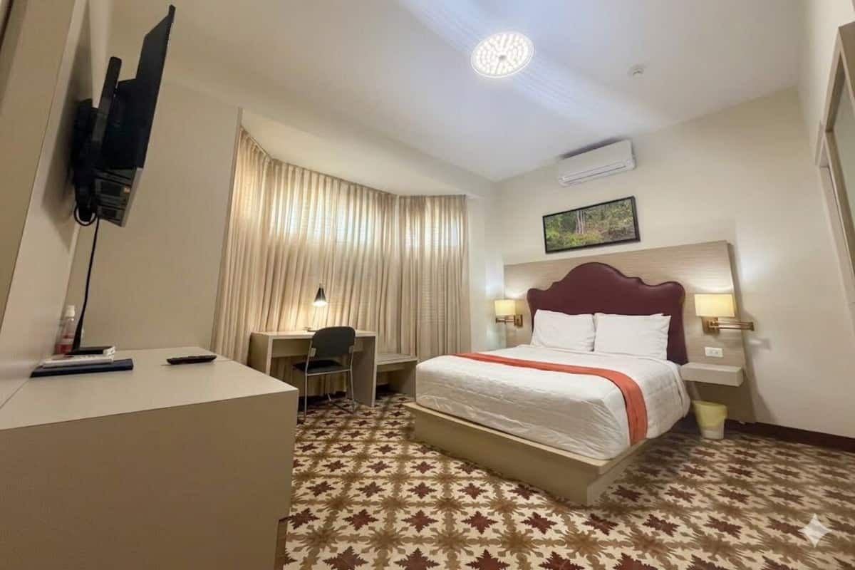 Una habitación de hotel con una cama, escritorio, silla y televisor sobre un soporte.