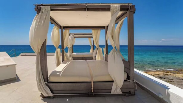 A four-poster canopy bed with beige bedding, positioned on a balcony overlooking the sea.