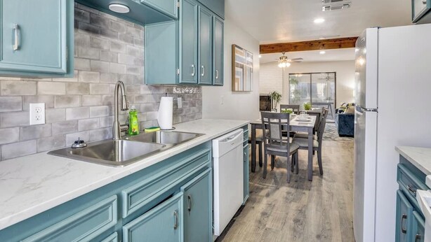 Une cuisine avec des meubles de couleur turquoise, un lave-vaisselle blanc et un coin repas comprenant une table et des chaises en bois.