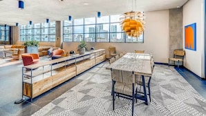A spacious, modern lounge area with a long wooden bench, a central table, and a hanging pendant light. Large windows provide views of the cityscape.