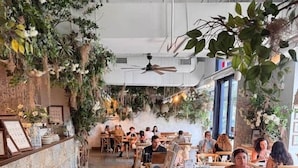 A restaurant with a long counter, multiple tables, and hanging plants.