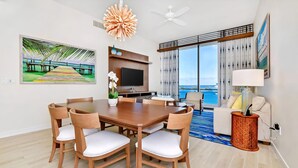 A modern hotel room with a dining area, a TV, a large window with a view of the sea, and a ceiling fan.