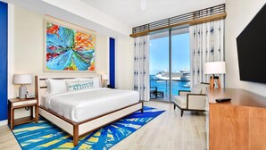 A hotel room with a bed, a desk, a chair, a large window with a view of a cruise ship, and a colorful abstract painting on the wall.