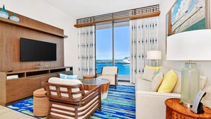 A modern hotel room with a large TV, comfortable seating, a view of the sea, and a balcony with curtains.
