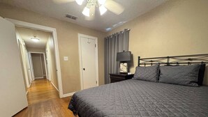 A bedroom with a large bed, a nightstand, a lamp, a ceiling fan, and a door leading to another room. - The Pedal Pad - 3 BED / 1 BATH @ central location (Wichita Falls)