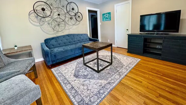 A living room with a blue sofa, a coffee table, a TV, and a wall-mounted bicycle art piece. - The Pedal Pad - 3 BED / 1 BATH @ central location (Wichita Falls)
