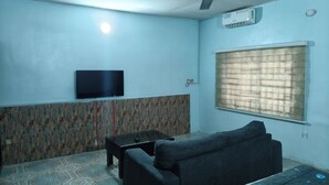 A living room with a flat-screen TV, a sofa, a coffee table, patterned wall, and a window with blinds. - Oasis Place (Port Harcourt)